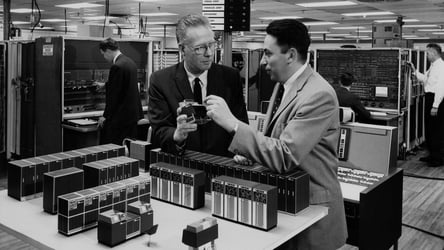 A black and white photo of two people talking about technology, surrounded by vintage hardware.